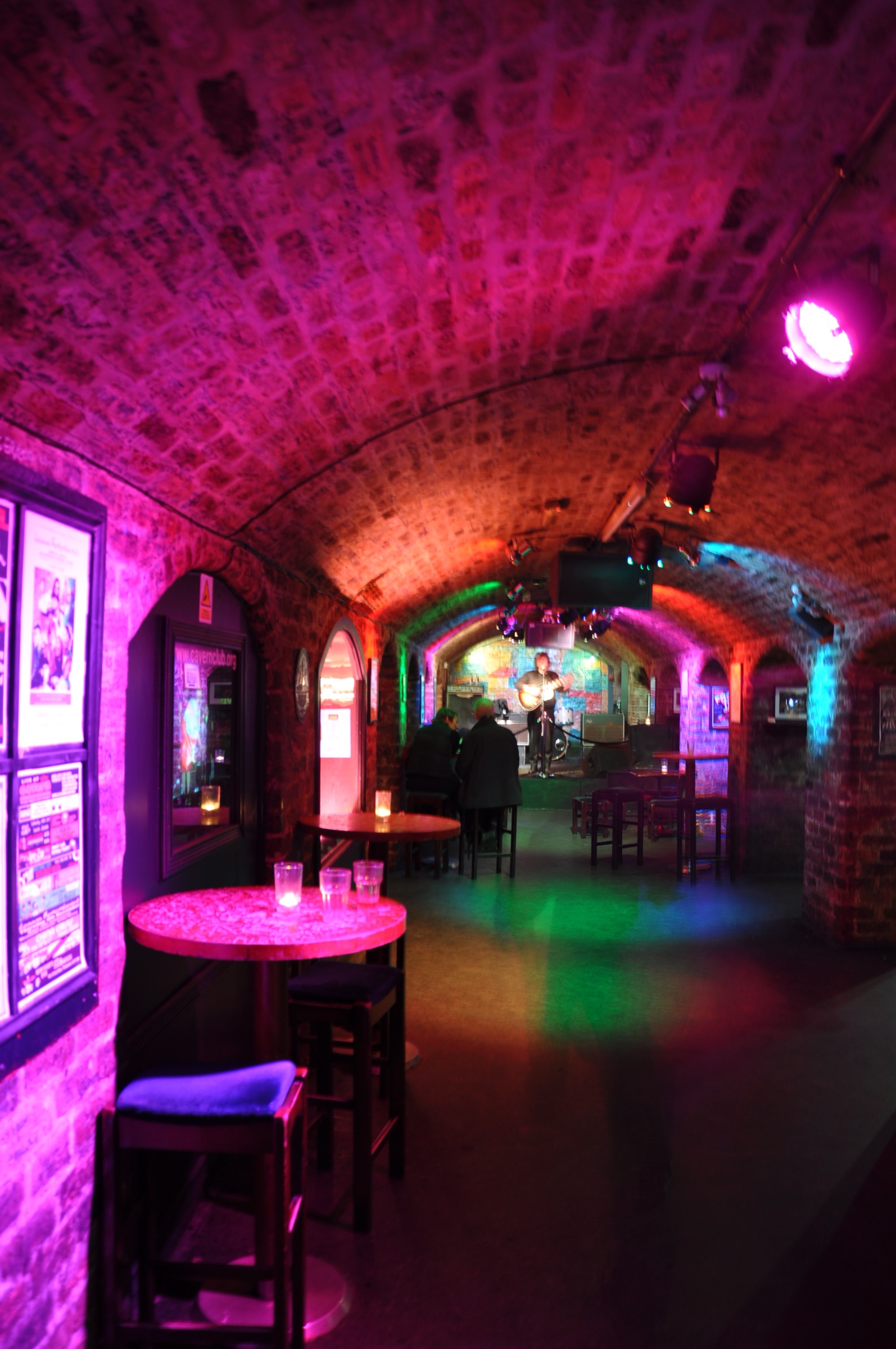 Brick-vaulted arches inside the rebuilt Cavern Club on Mathew Street, Liverpool, lit in pink and green, with a musician on the small stage at the far end