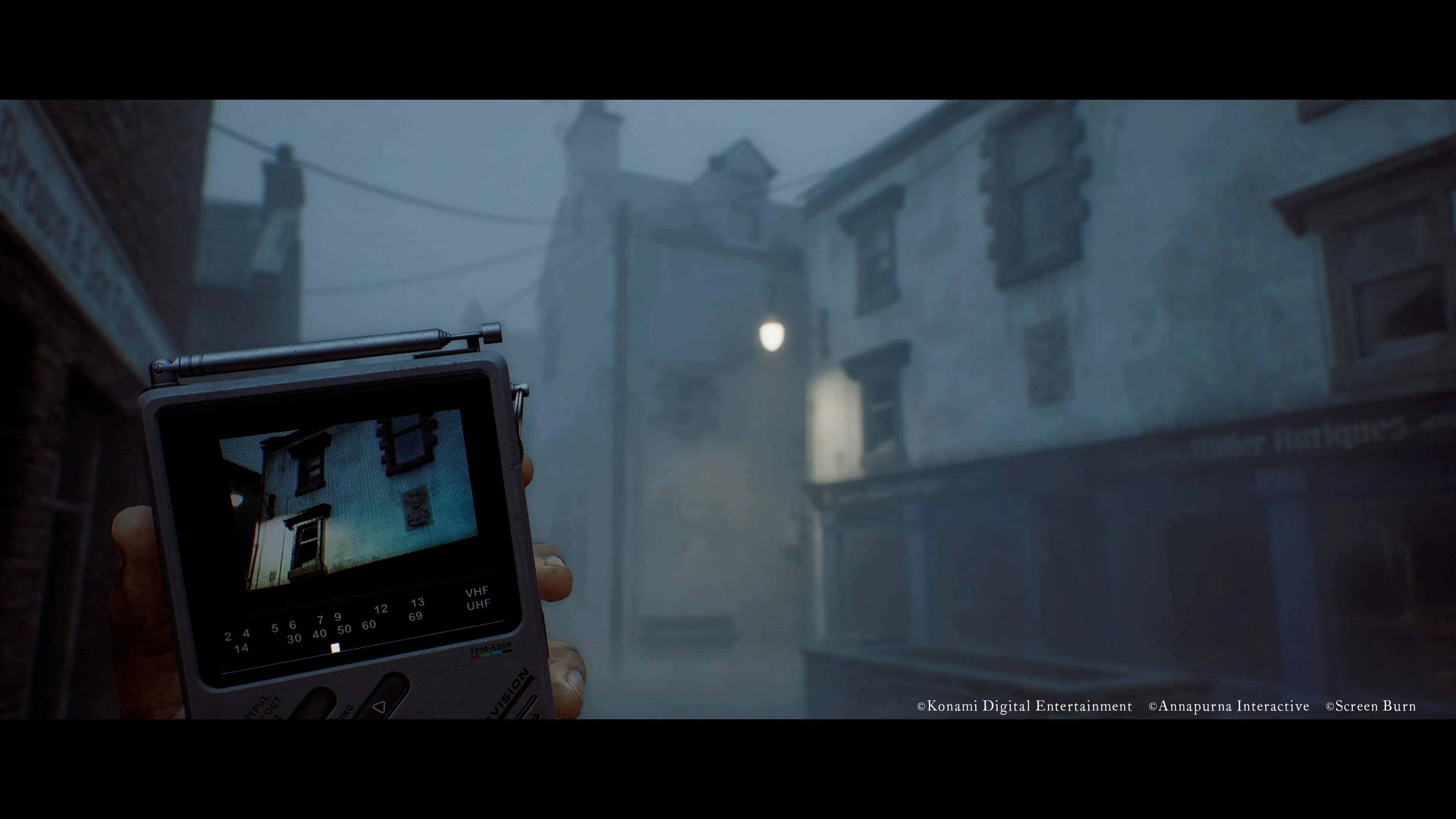 A hand holds up a handheld analog tuner device showing a grainy image, against a foggy Scottish street of stone buildings, from Silent Hill: Townfall