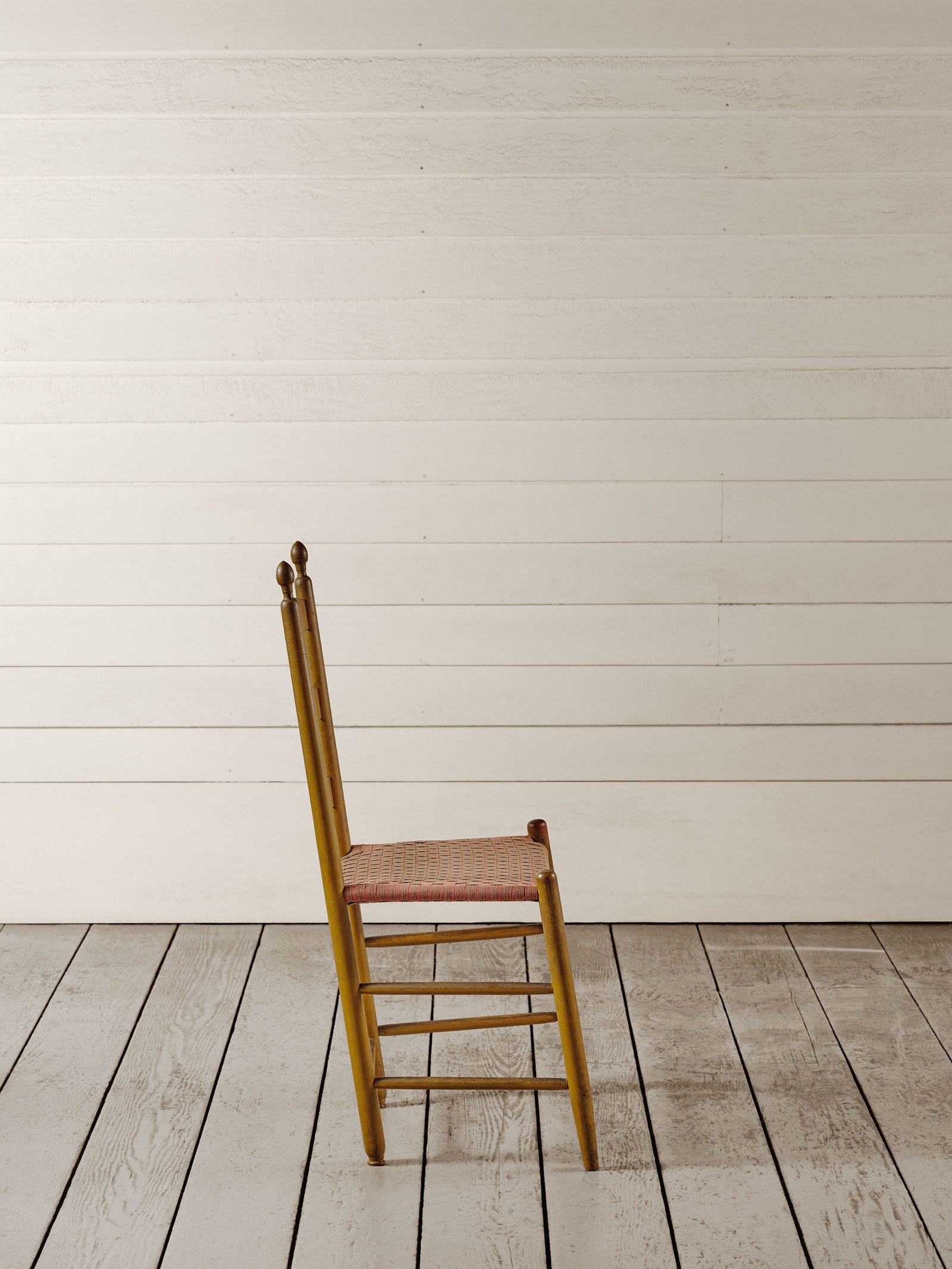 A plain yellow-painted Shaker ladder-back side chair with a woven seat, shown in profile against white clapboard walls and a whitewashed plank floor