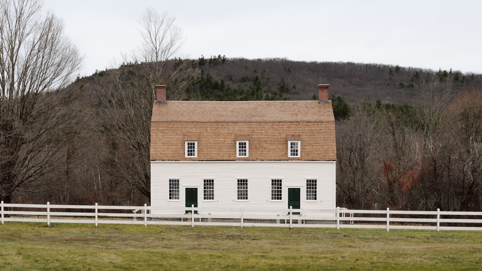The white clapboard 1793 Shaker Meetinghouse with a gambrel roof and twin chimneys, behind a white rail fence at the foot of a wooded hill