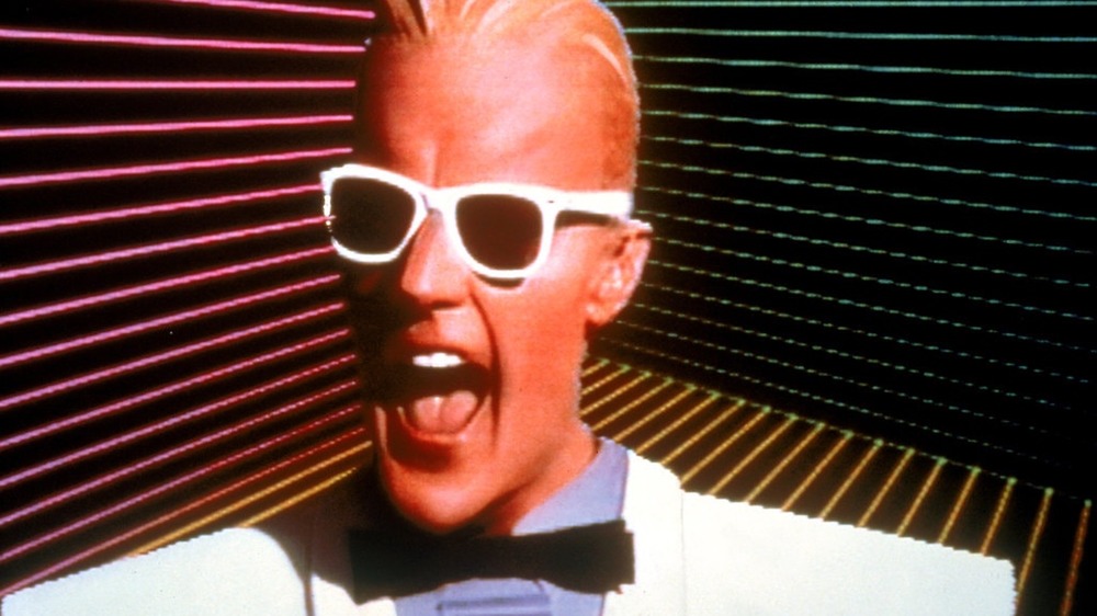 Matt Frewer as Max Headroom wearing white Wayfarer sunglasses and a bow tie, mouth open mid-line, against a colorful striped backdrop