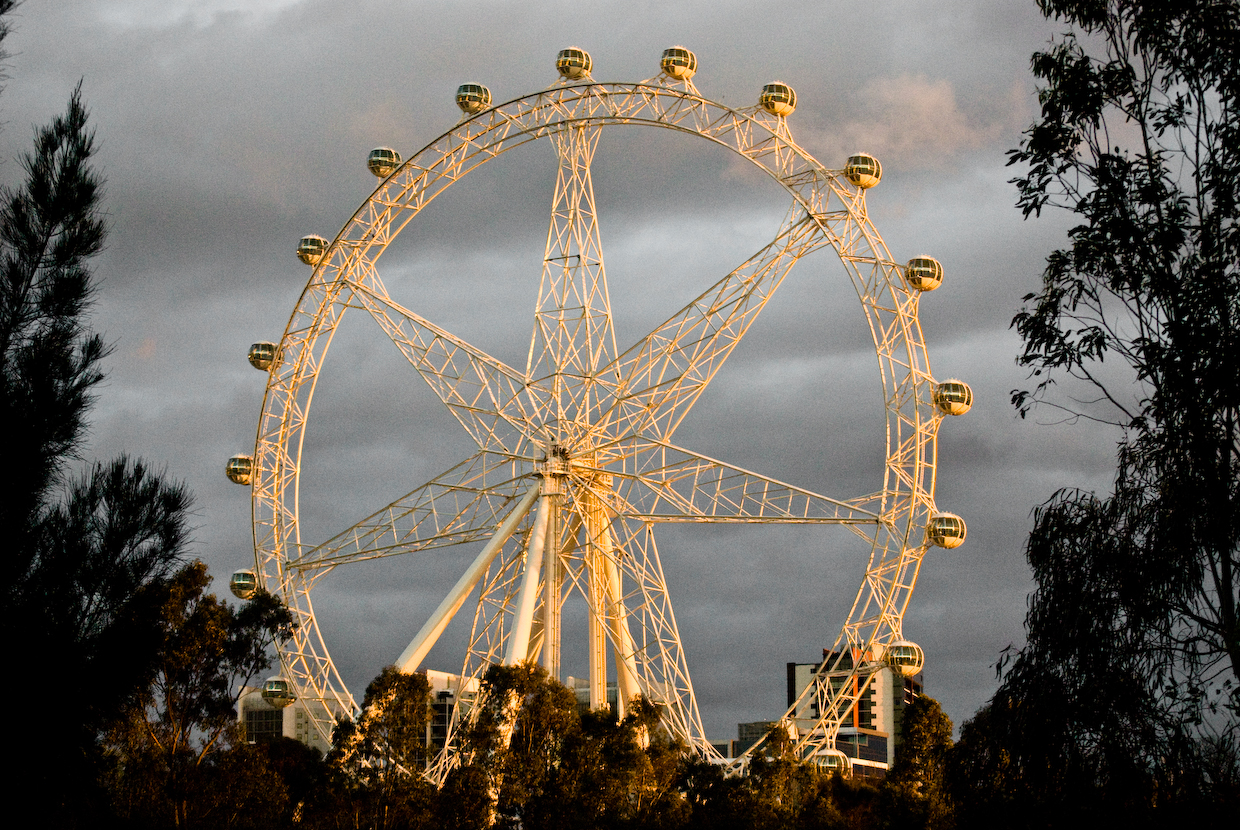 The Southern Star observation wheel in Melbourne at dusk, its white seven-spoke frame and round cabins lit gold against a grey sky, framed by dark trees