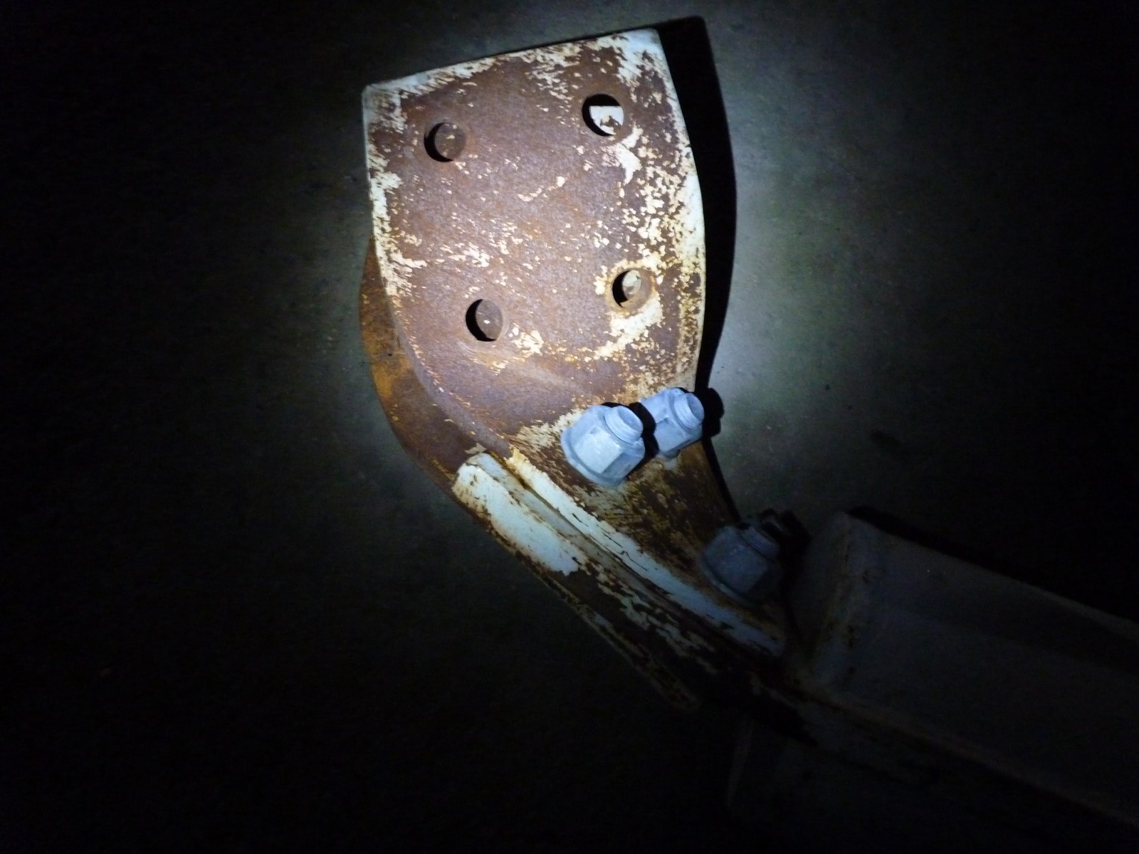 A rusted, buckled steel joint plate with bolt holes and white paint flaking off, lit by a single torch against a black background
