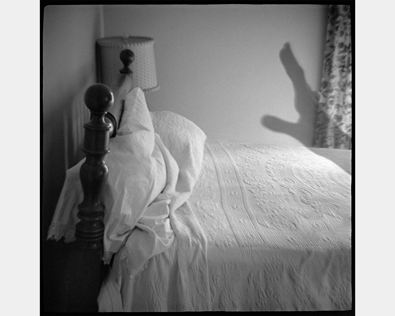 A black-and-white photograph of a made bed with a dark wooden bedpost, a lamp and a puffed pillow, and on the wall behind it the long shadow of a hand
