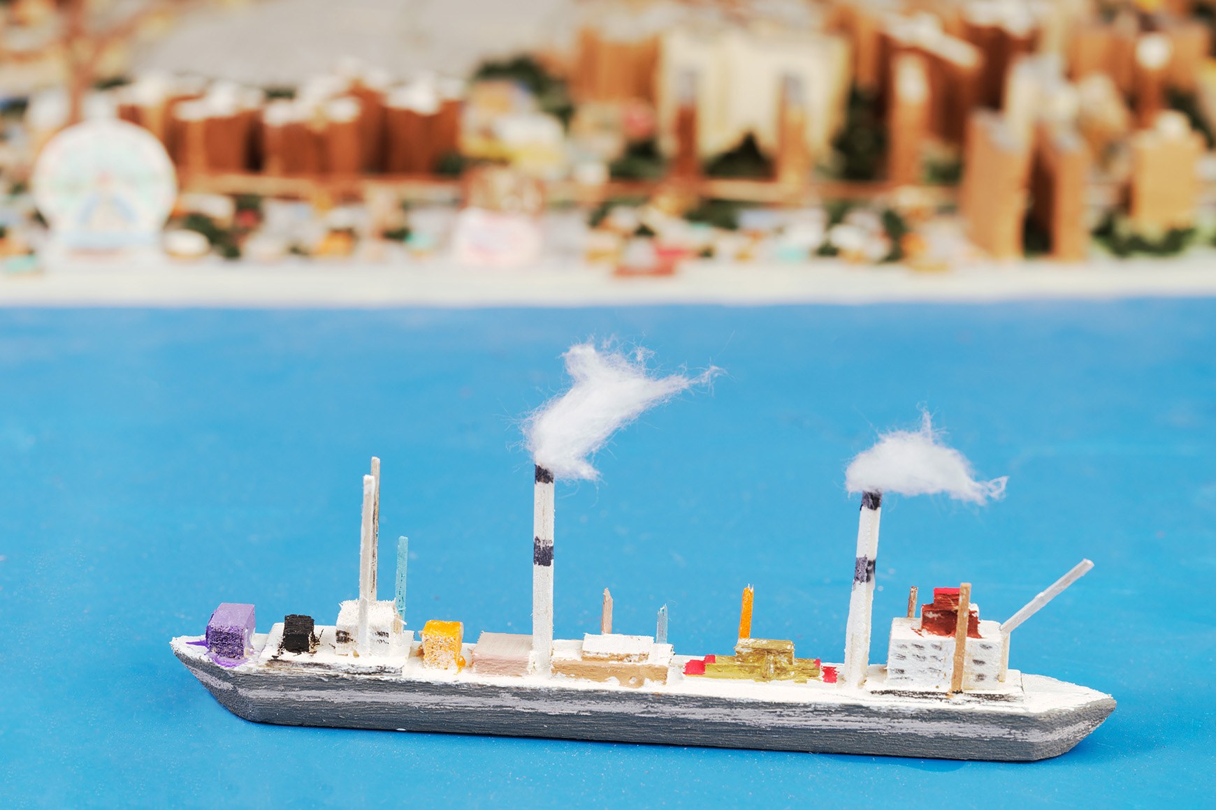 A small handmade boat with two smokestacks and cotton-wool smoke, sitting on blue painted water in the model