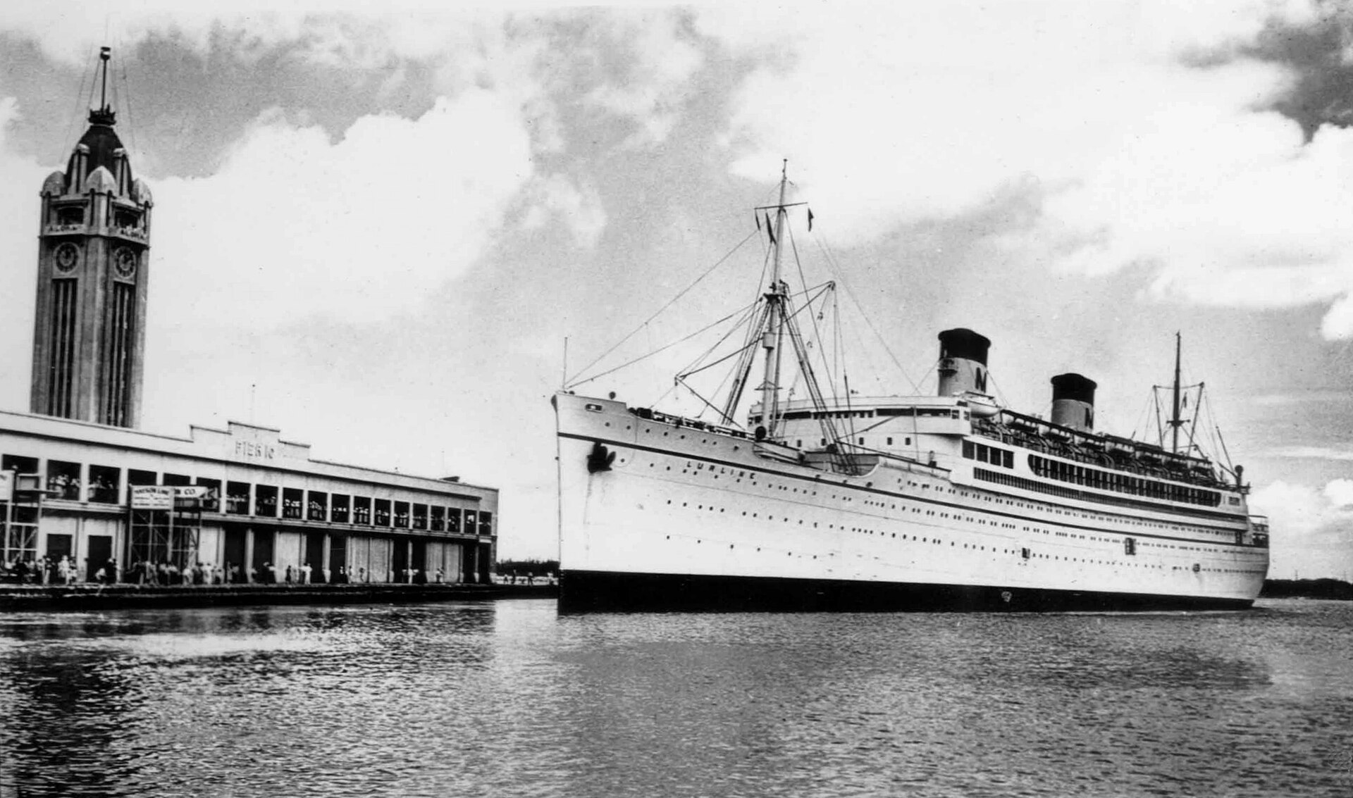 Black and white photo of the liner SS Lurline alongside a pier at Honolulu with the Aloha Tower rising behind the pier shed