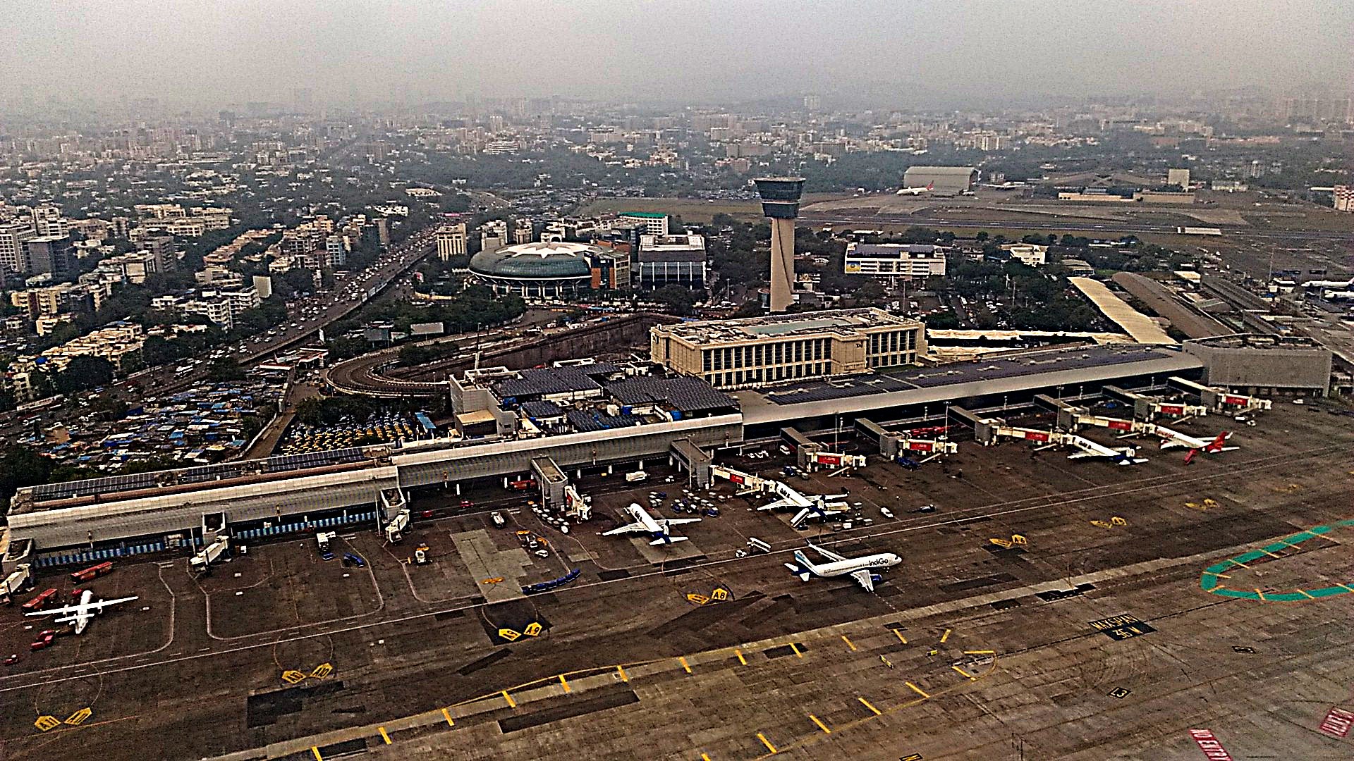 Aerial view of Mumbai airport's Terminal 1: a long low terminal with jet bridges and parked planes, the city spreading behind it under haze