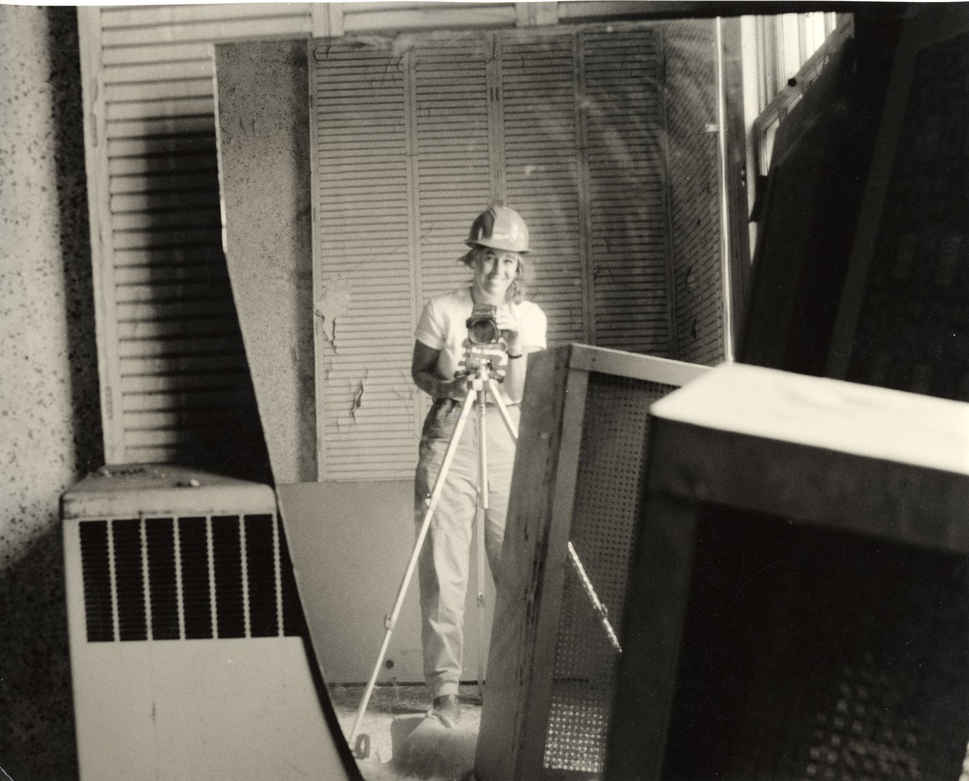Black-and-white self-portrait: a woman in a hard hat behind a camera on a tripod, reflected in a broken mirror among stacked radiator covers and shutters