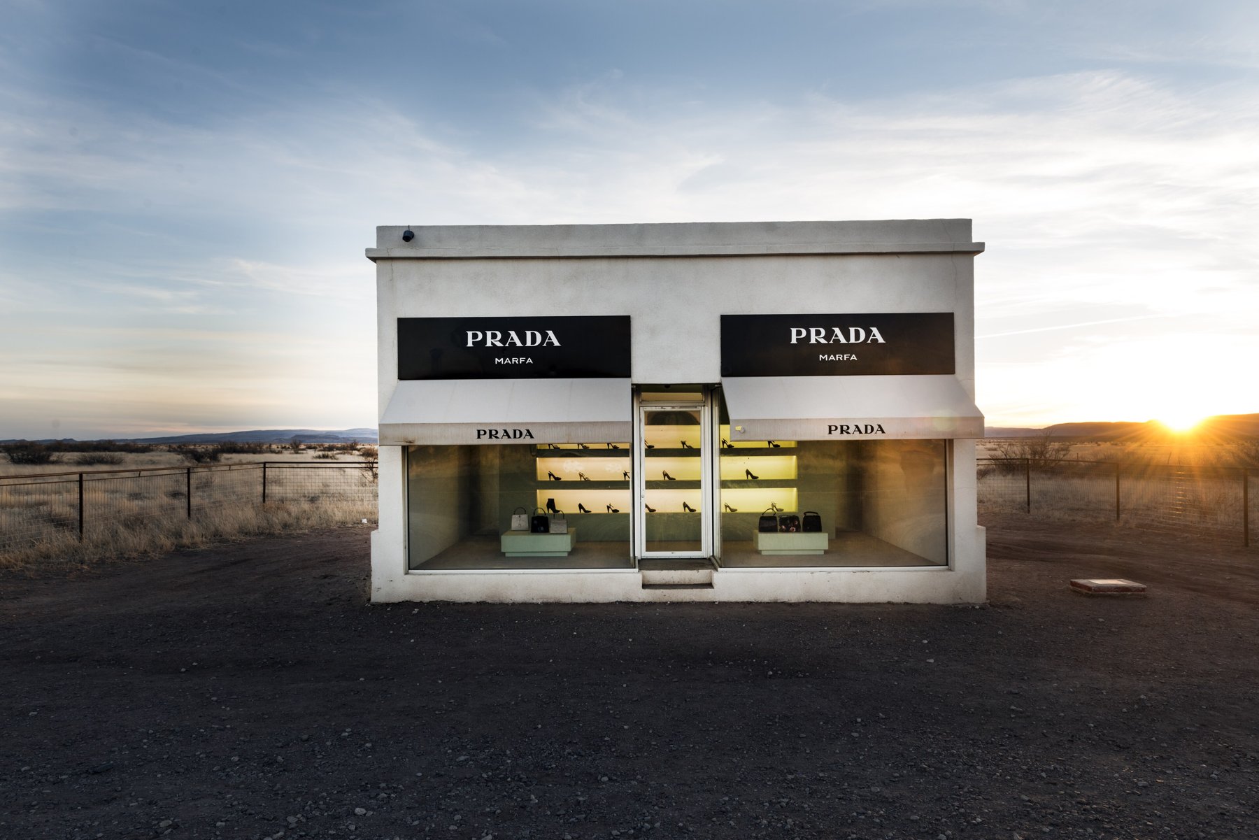 Prada Marfa at sunset: a small white boutique with PRADA awnings and shoes in the window, alone on gravel in the West Texas desert