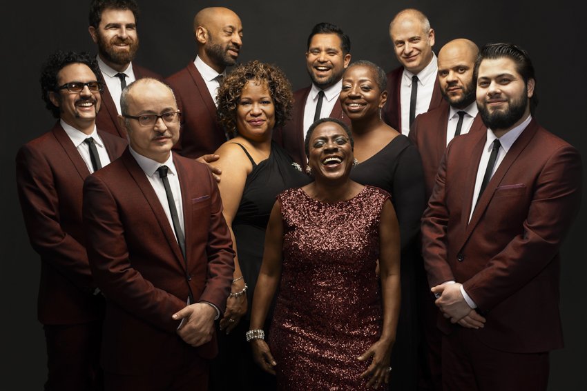 Sharon Jones in a sequinned dress, smiling, surrounded by the Dap-Kings in matching burgundy suits