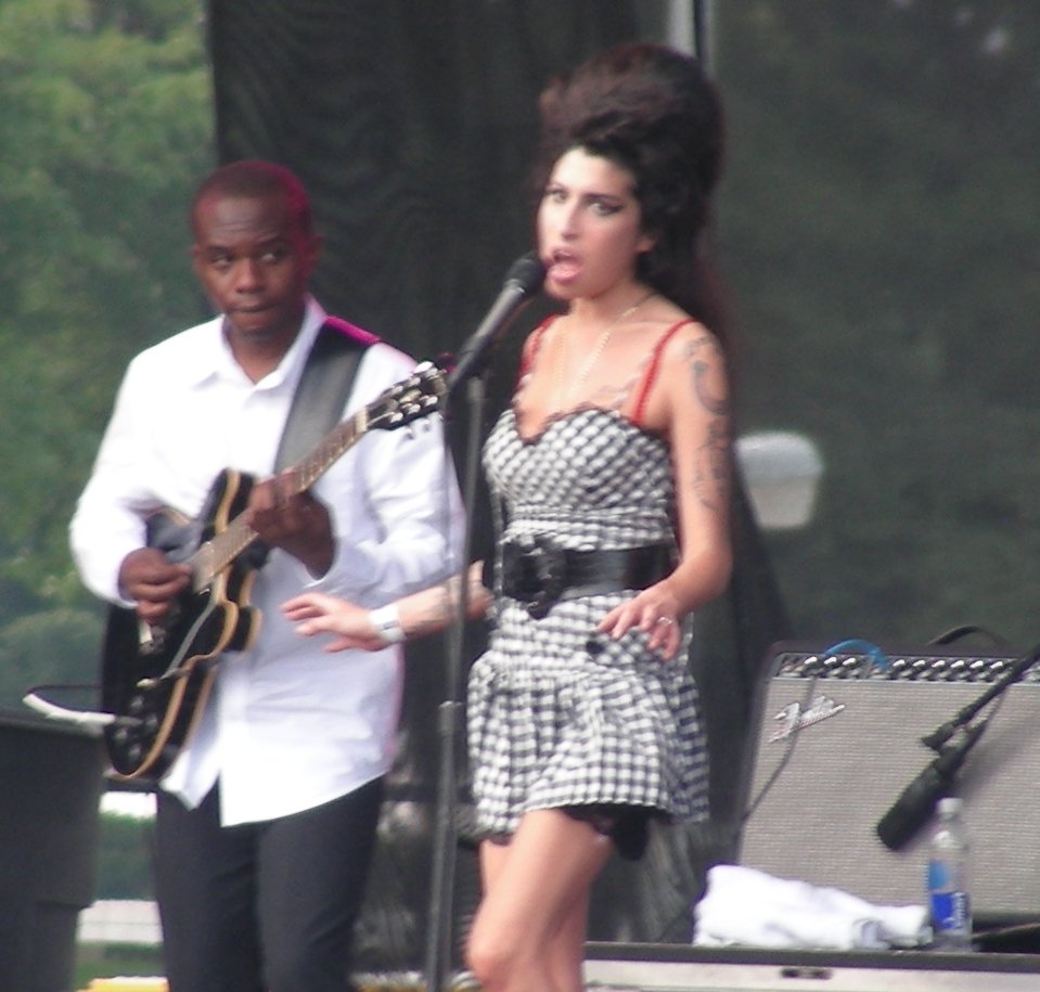 Amy Winehouse singing on an outdoor festival stage in a checked dress, a guitarist in a white shirt beside her
