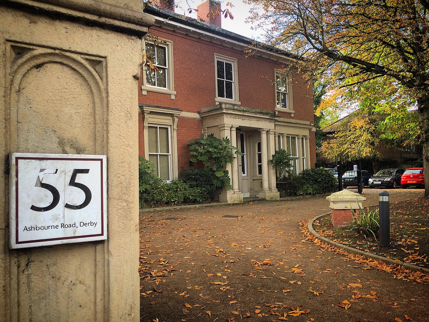 A brick Victorian house behind a stone gatepost with a sign reading 55 Ashbourne Road, Derby, autumn leaves on the drive