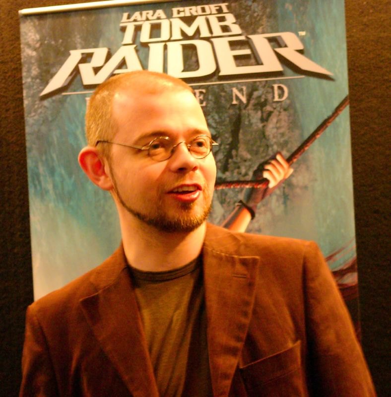 Toby Gard, in glasses and a brown corduroy jacket, in front of a Tomb Raider: Legend banner