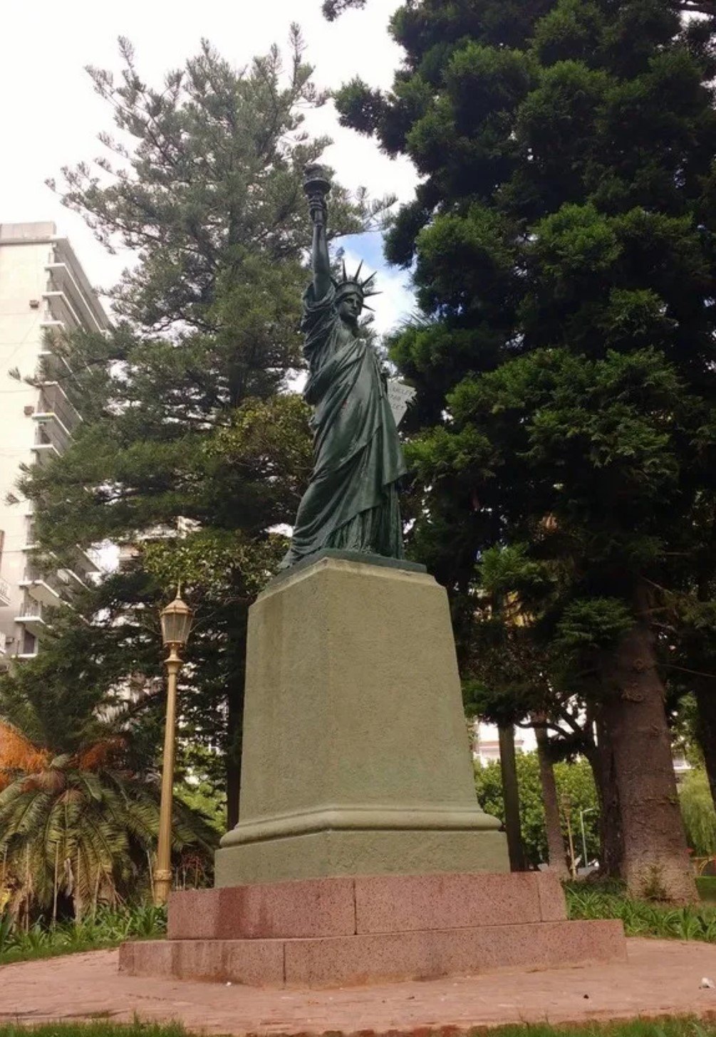 A small green Statue of Liberty on a tall stone pedestal among pines and palms in a Buenos Aires park, apartment blocks behind