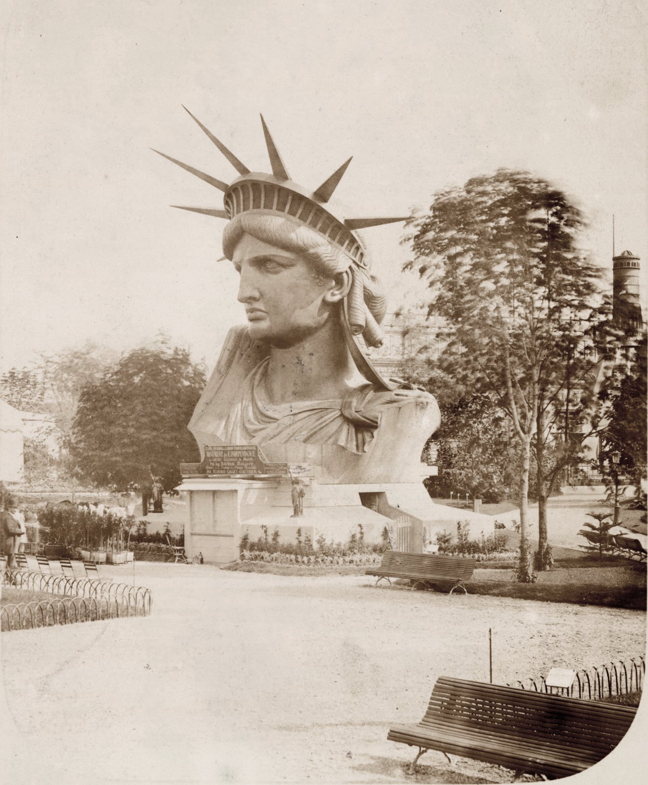 Sepia photograph of the Statue of Liberty's giant head and crown standing on the ground in a Paris park, with trees and a bench around it