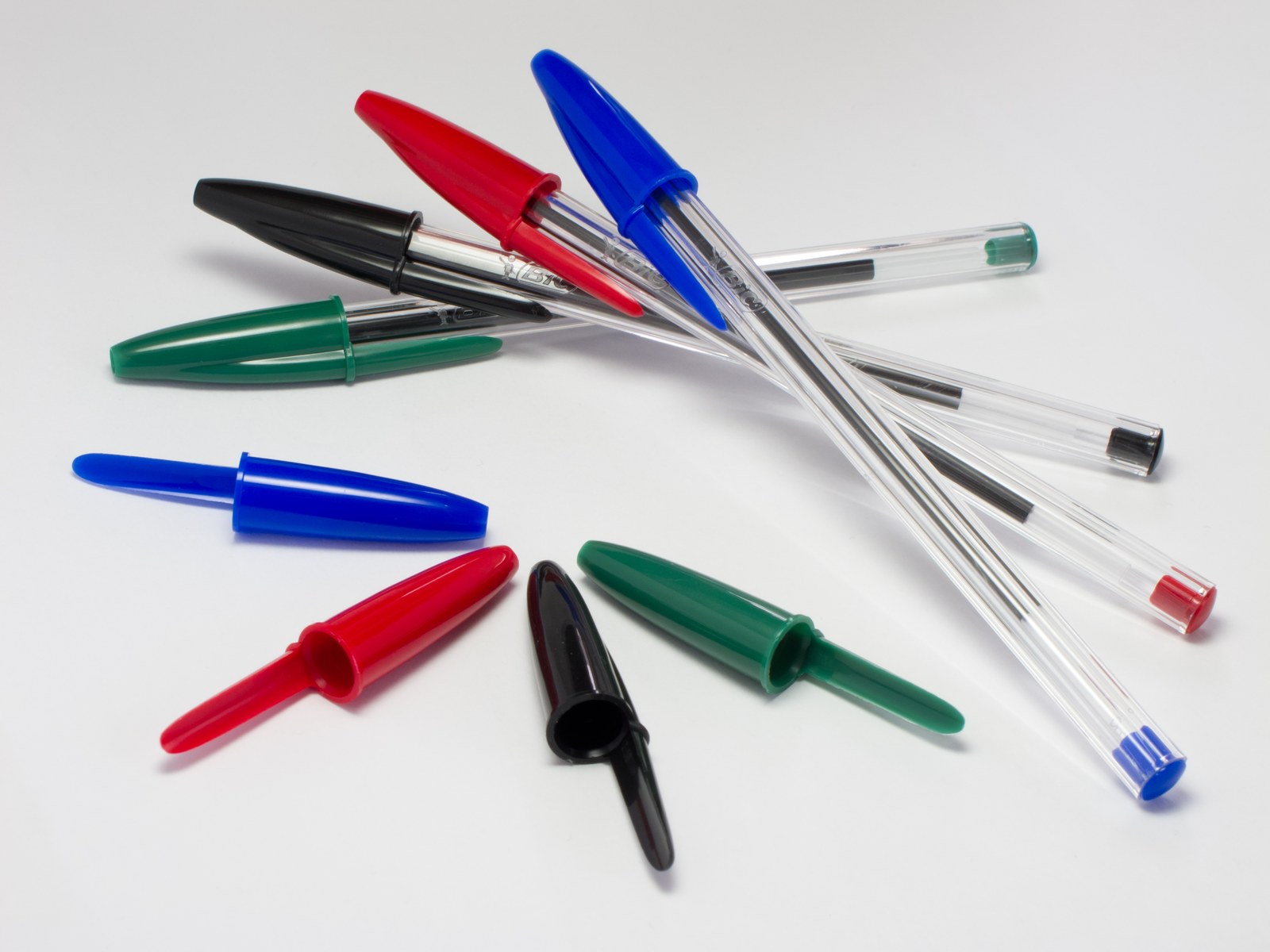 Four Bic Cristal pens, blue, red, black and green, with their caps scattered on a white surface